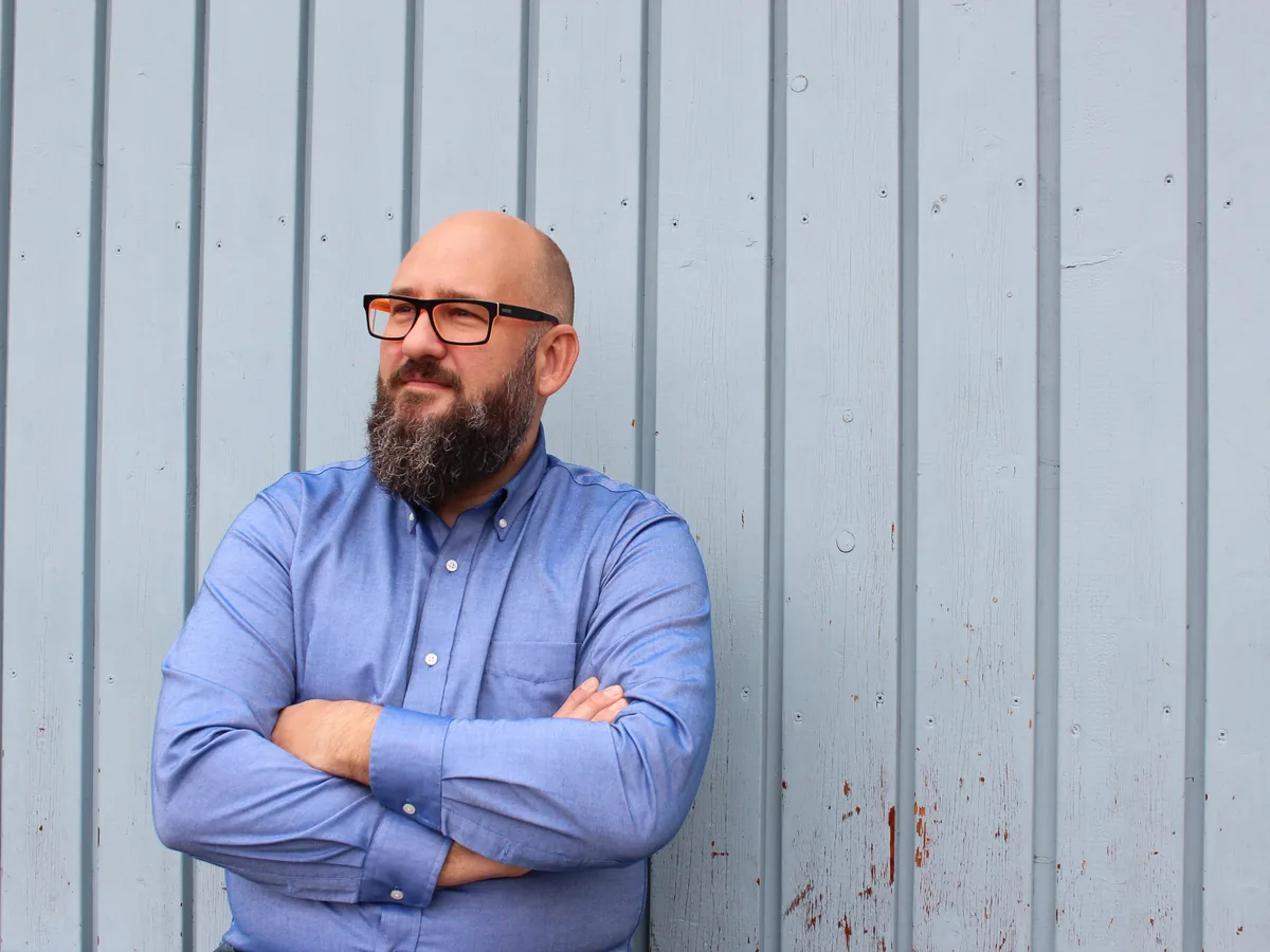 Willi Rauch mit verschränkten Armen vor einer hellblauen Holzwand